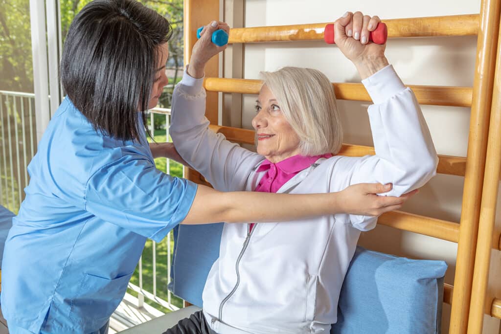A caregiver assists an elderly woman with strength exercises using dumbbells at a wooden stall bar. The scene is encouraging and supportive.