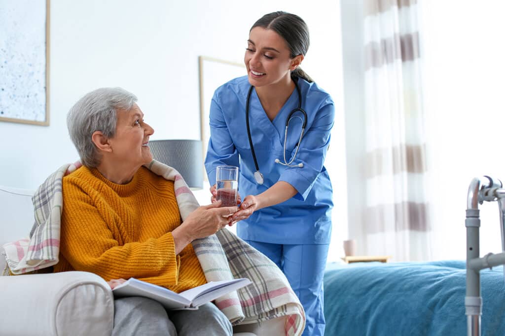 A nurse in blue scrubs, smiling, hands a glass of water to an elderly woman in a mustard sweater.