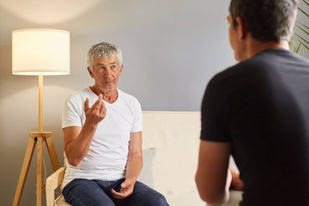 Older man in a white shirt sits on a sofa, gesturing expressively while speaking to a person in a black shirt.