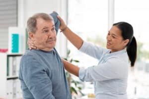 A physical therapist is helping an elderly man in a blue shirt stretch his arm.