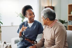 A nurse is holding a tablet while sitting on a couch, talking with an elderly man.